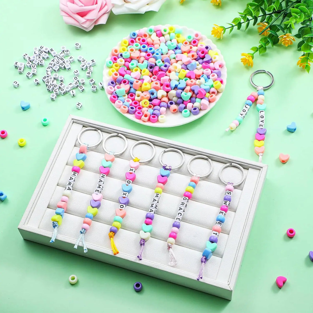 A white tray holds several heart-shaped beaded keychains with letter charms in various pastel colors.