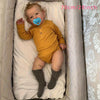 A baby doll dressed in a mustard yellow outfit with gray socks and a blue pacifier is lying in a padded enclosure.