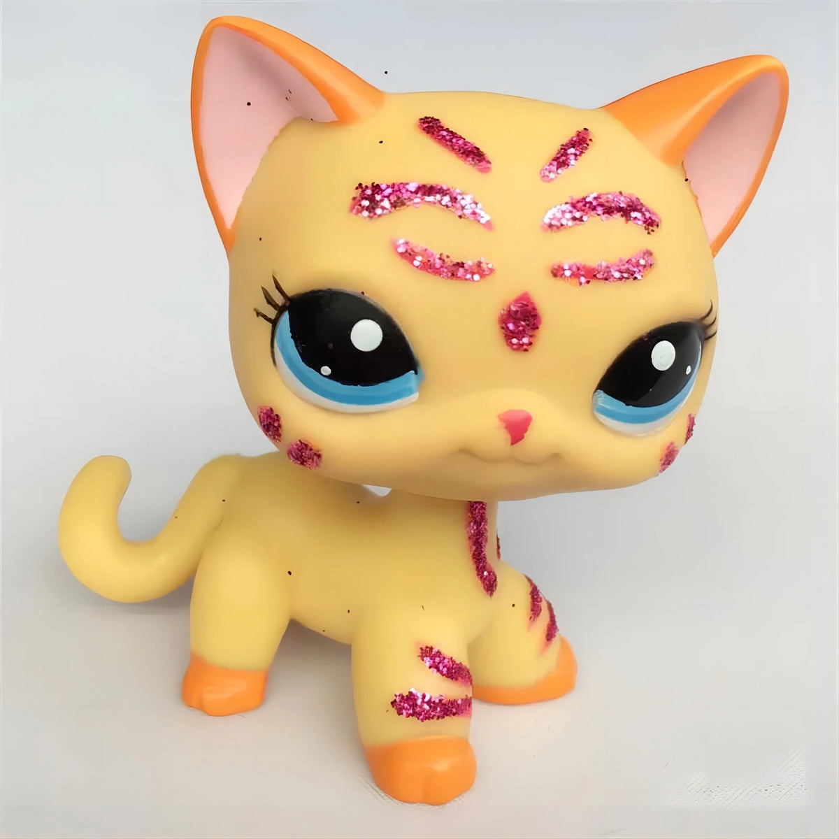A yellow Littlest Pet Shop style cat figure with pink glitter accents and large blue eyes.