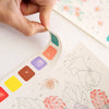 A hand is pointing at a color palette section in a creative watercolor painting book for kids featuring fairy tale and animal