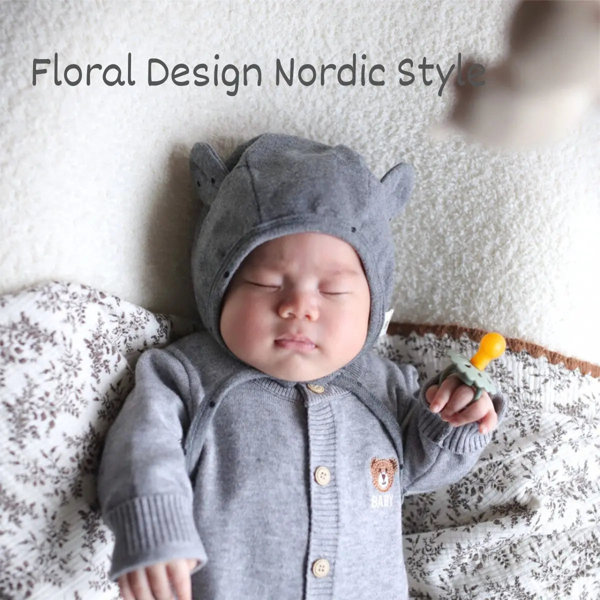 A newborn baby wearing a gray hooded outfit and holding a natural latex pacifier on a white blanket.