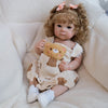 A baby doll with hand-rooted curly hair wearing a light brown outfit and sitting in a fluffy white blanket.
