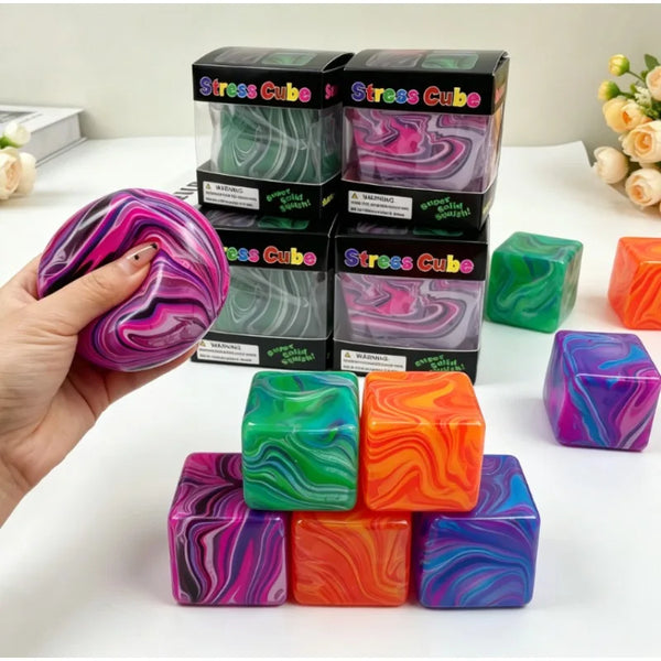 A hand holds a purple and pink stress cube next to several packaged green, pink, and orange stress cubes.