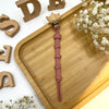 A pink baby pacifier clip with a burlap flower design rests on a wooden tray surrounded by wooden letters.