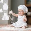 A baby wearing a white chef apron and hat is playing with flour on a wooden surface.