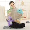 A woman holds a green plush dinosaur and purple and orange plush toys on a white rug.