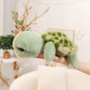 A hand holds a soft, light green Big Eyes Tortoise plush pillow with textured skin and leafy patterns.