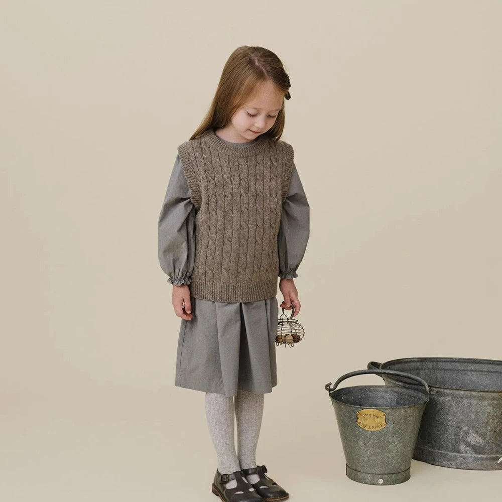 A young girl wearing a beige knit sweater and gray pleated skirt with matching tights.