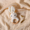 A handmade white crochet bunny rattle set resting on a light beige linen fabric with gold leaves.