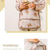 A baby wearing a light beige waterproof long sleeve feeding bib decorated with small bear patterns.