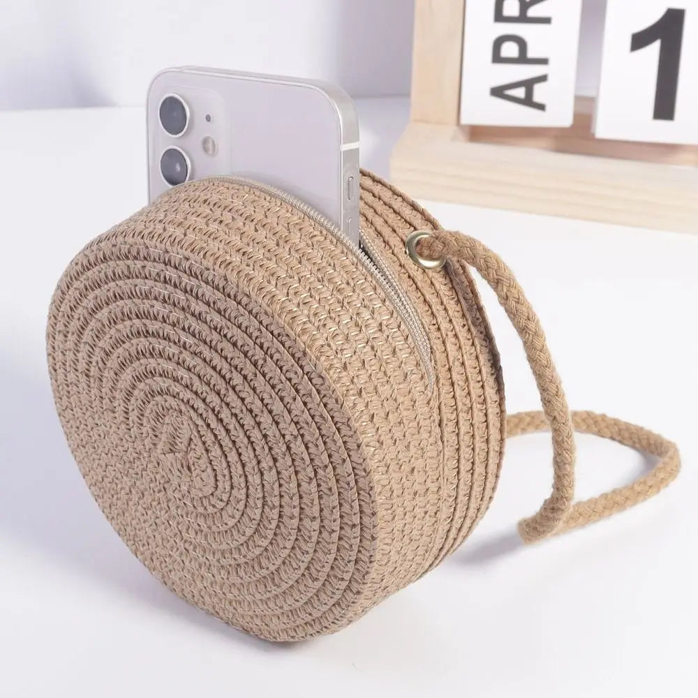 A beige handwoven straw crossbody bag with a zipper and lace bow next to an iPhone.