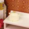 A miniature cream-colored copper soup pot with a lid displayed on a white shelf against a patterned background.