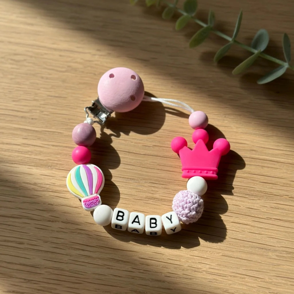 A pink silicone pacifier clip bracelet featuring various colored beads and a crown charm on a wooden surface.