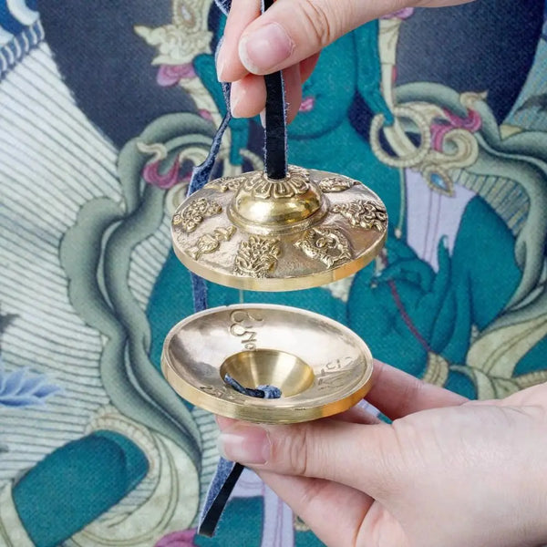 A hand holds a handcrafted brass Tingsha cymbal with eight auspicious symbols against a patterned fabric.