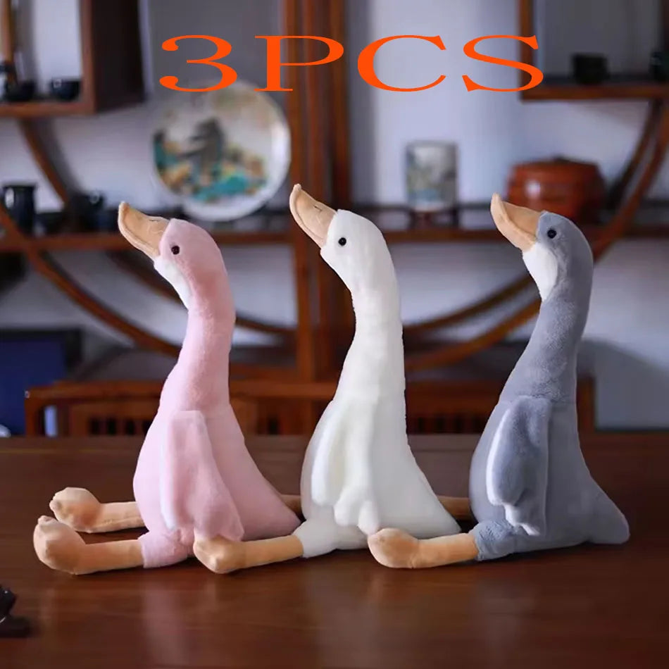 Three plush toys of long-necked geese in pink, white, and gray colors displayed on a wooden surface.