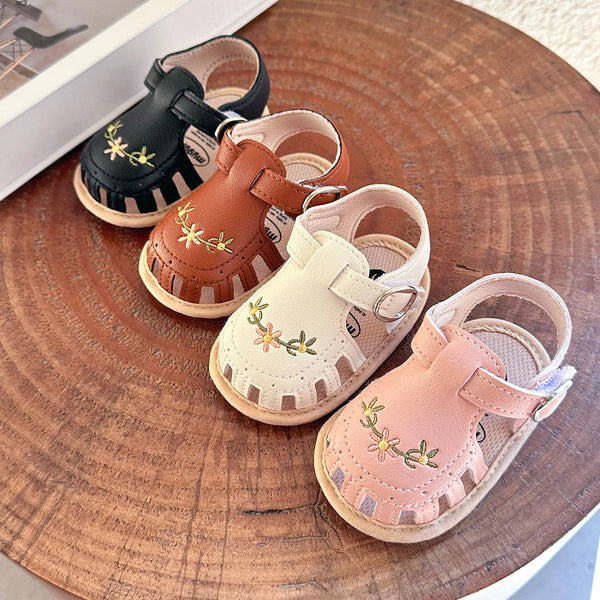 Three pairs of infant and toddler unisex sandals in brown, tan, and pink leather with floral accents displayed on a wooden su