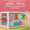 A hand holds a white Montessori shape sorting cube with colorful plastic shapes and elastic bands on a green surface.