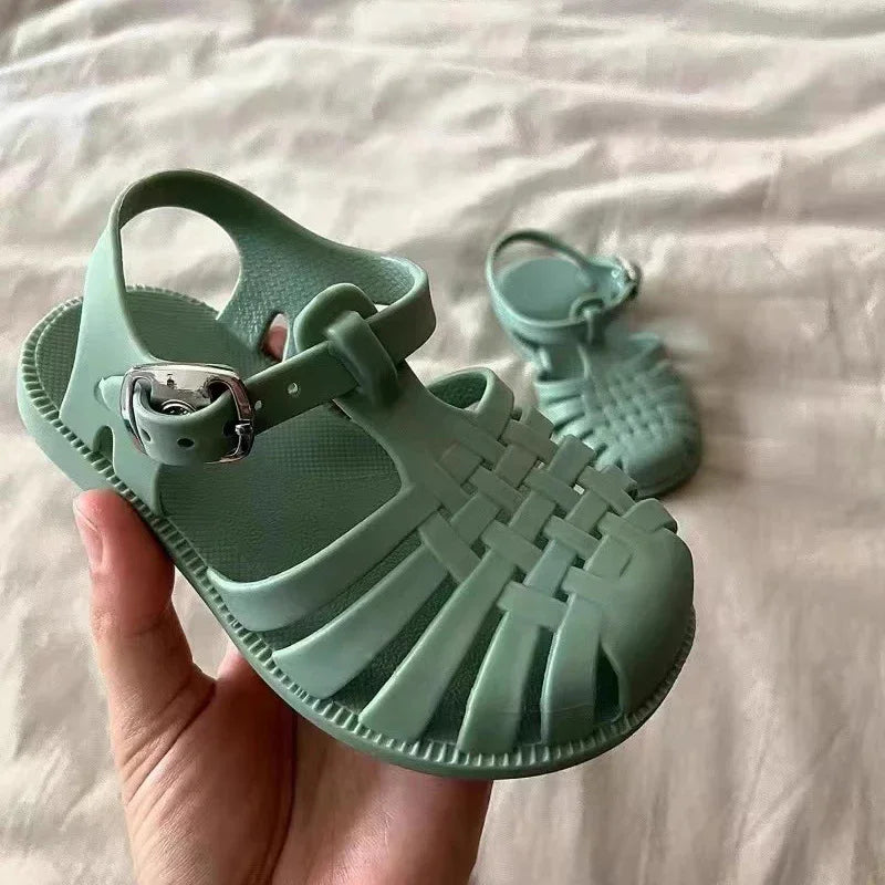 A hand holds a pair of mint green girls' gladiator jelly sandals with non-slip soles.