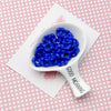 A white ceramic spoon holds a cluster of bright blue acrylic beads with floral edges on a pink patterned surface.