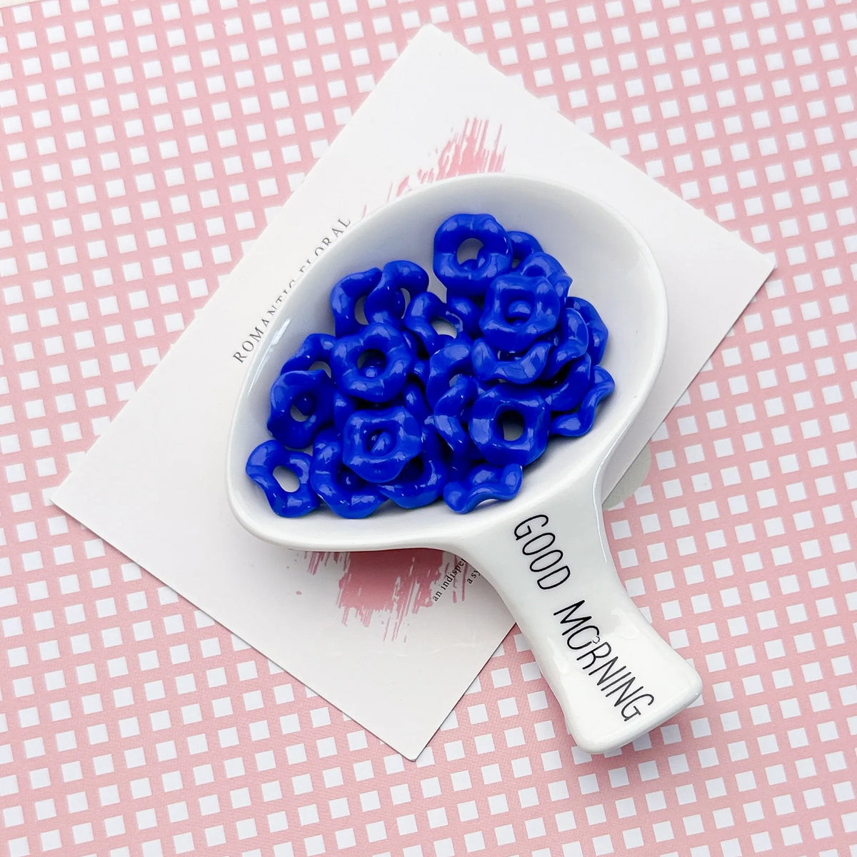 A white ceramic spoon holds a cluster of bright blue acrylic beads with floral edges on a pink patterned surface.
