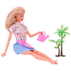 A blonde doll wearing a floral top and blue skirt is shown with a pink watering can and a green palm plant accessory.