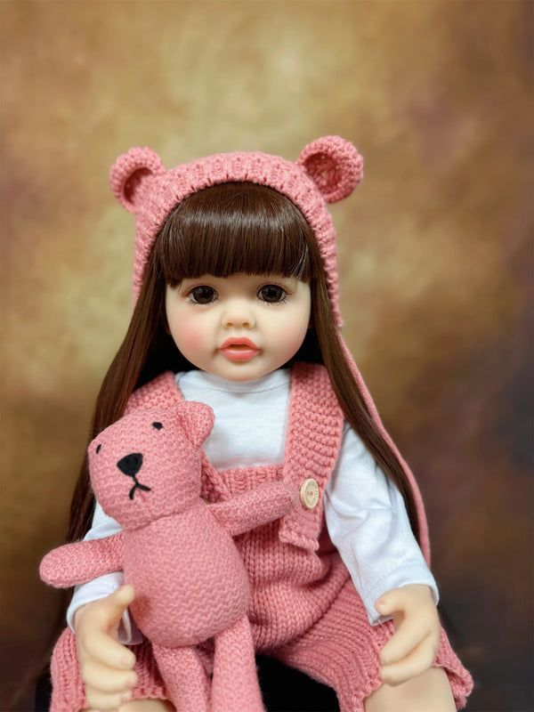 A realistic silicone baby doll with long dark hair wearing a pink bear hat and a matching pink knitted outfit.