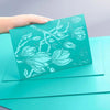 A hand displays a teal rubber plate with an engraved silhouette of leaves and branches, showing a sculpture effect display.