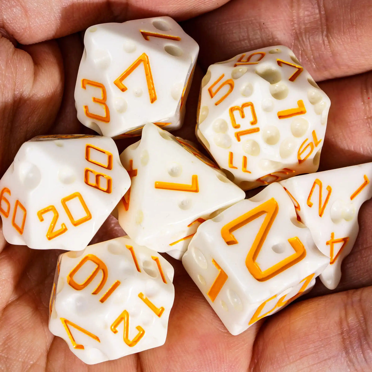 A hand holds seven white polyhedral dice with orange numbers, suggesting a cheese-themed set.