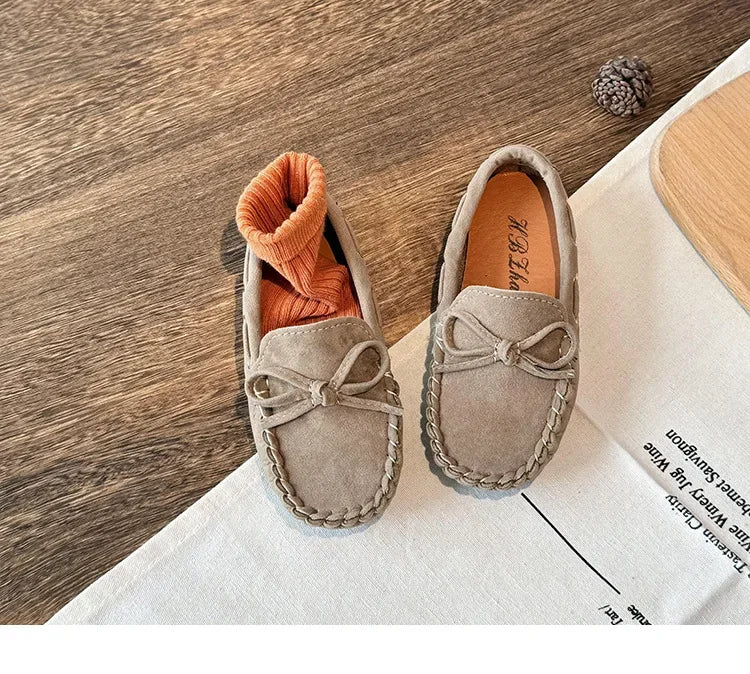 A pair of taupe slip-on moccasins with orange lining and bow details resting on a white cloth.