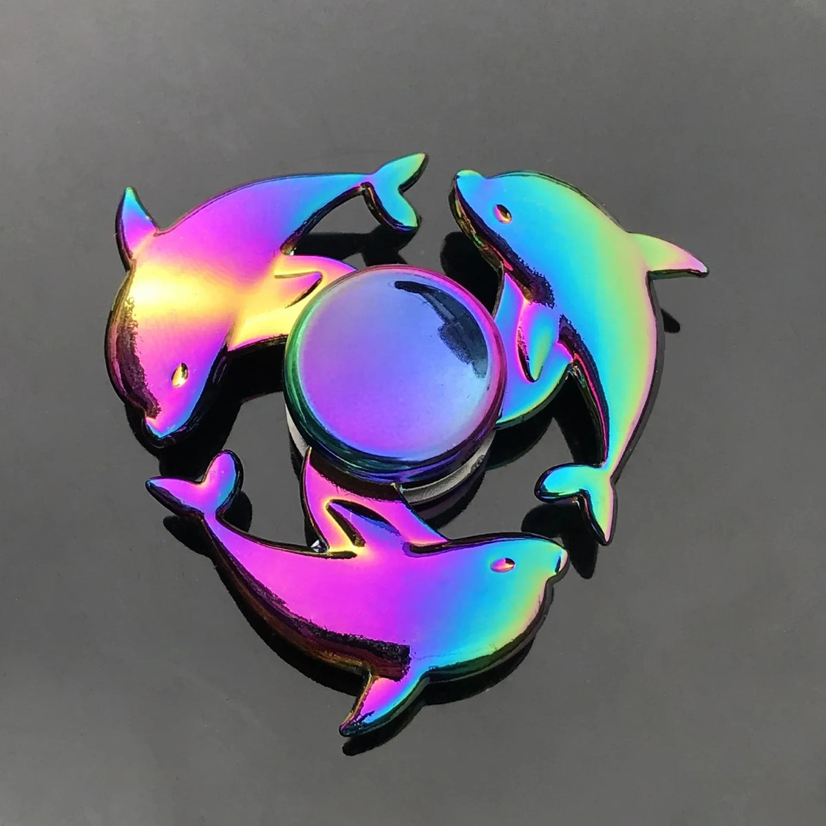 A rainbow metal fidget spinner featuring three stylized fish shapes rotating on a dark gray surface.