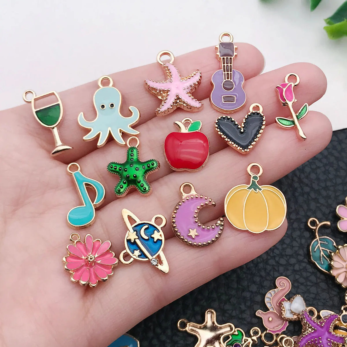 A hand holds various enamel alloy flower charms in pink, green, purple, and gold colors for DIY jewelry making.