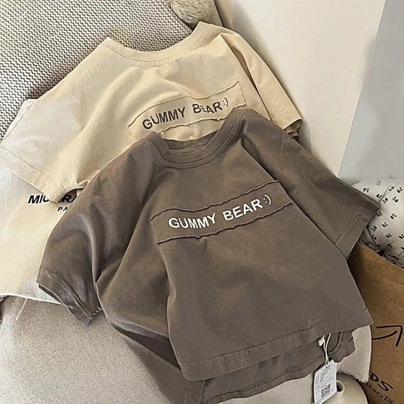 A close-up of a taupe short-sleeve t-shirt with the text GUMMY BEAR printed on the chest, lying on a light colored shirt.