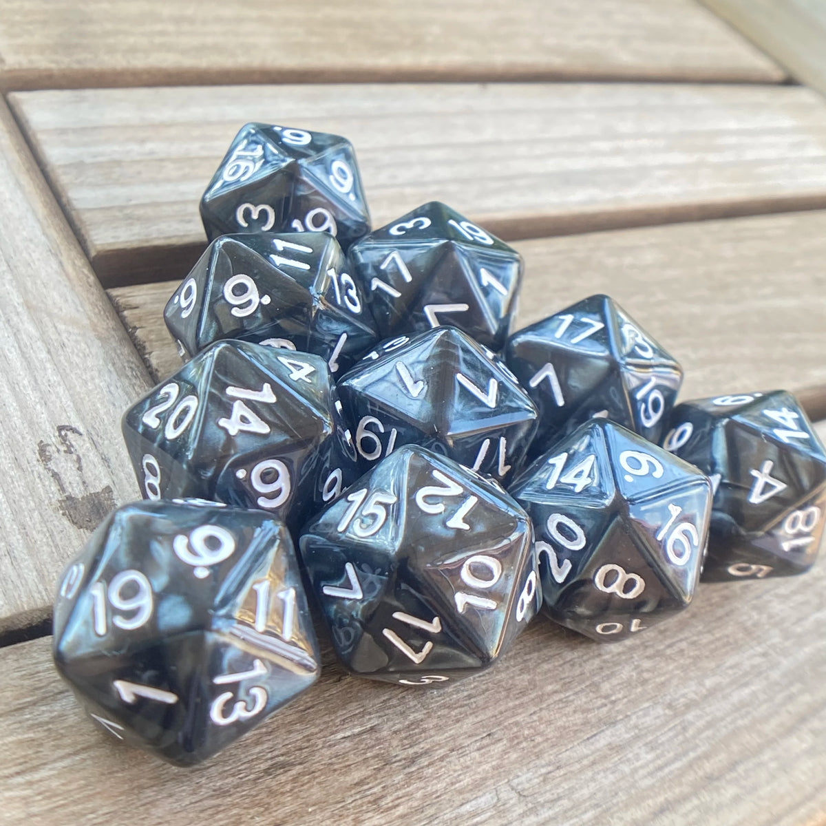 A pile of ten black and clear pearl acrylic D20 dice resting on a wooden surface.