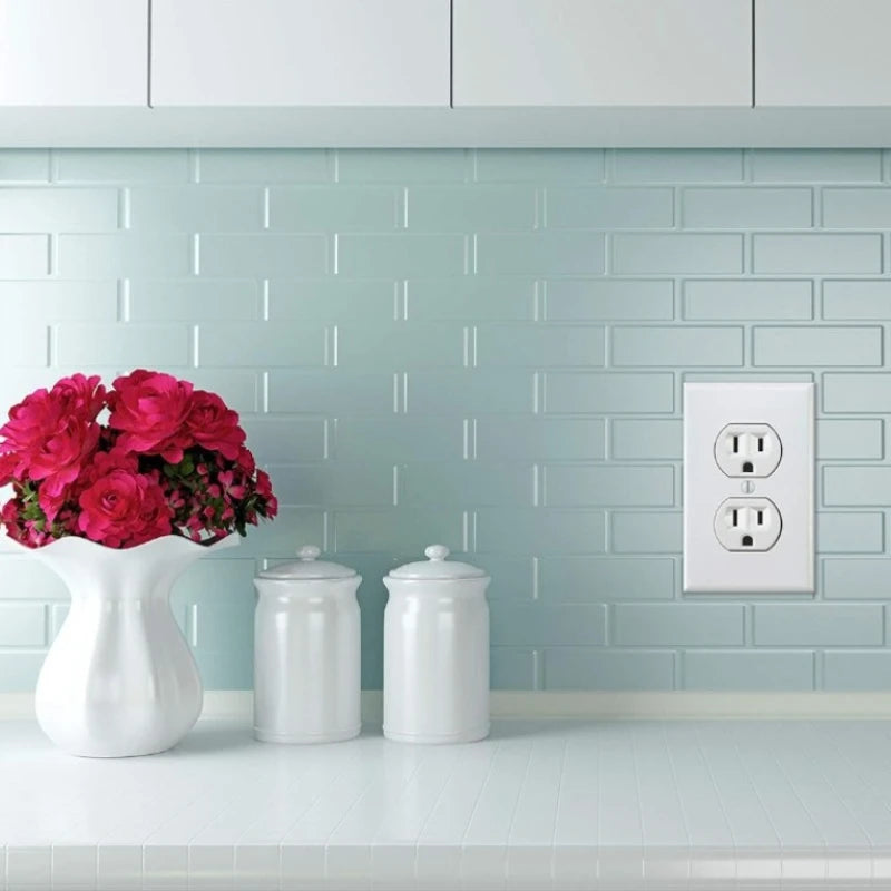 A white ceramic vase with pink roses sits on a light countertop in front of a light blue tiled wall featuring a white electri