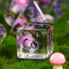A clear polyhedral die with a pink mushroom design rests on green grass.