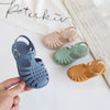 A hand holds a pair of girls' gladiator jelly sandals in various pastel colors, including blue, pink, green, and orange.