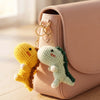 A handmade crochet dinosaur keychain with yellow and green colors hanging from a pink leather handbag.
