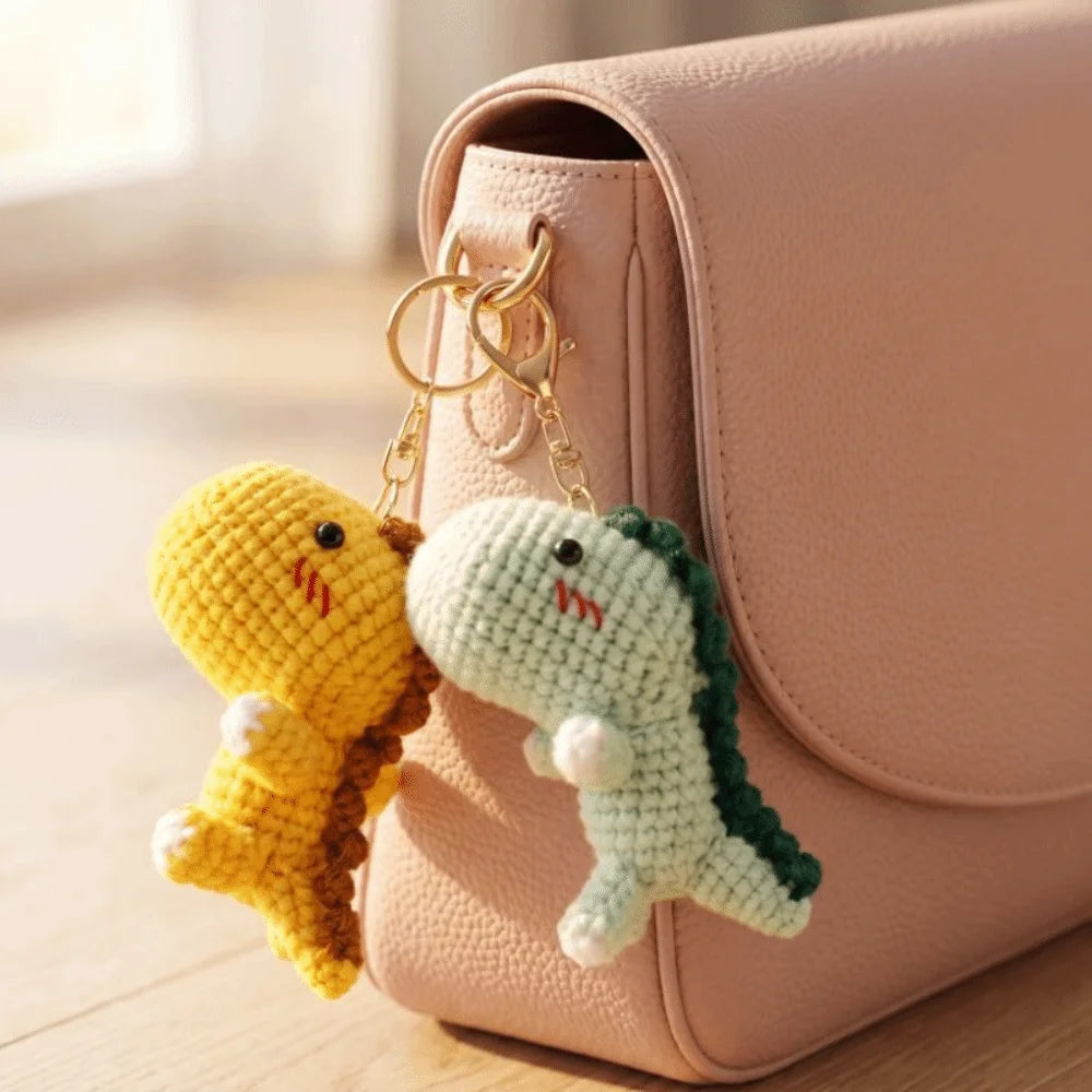 A handmade crochet dinosaur keychain with yellow and green colors hanging from a pink leather handbag.