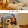 A young woman is sitting on a white sofa with a giant gray plush teddy bear, looking at her phone.
