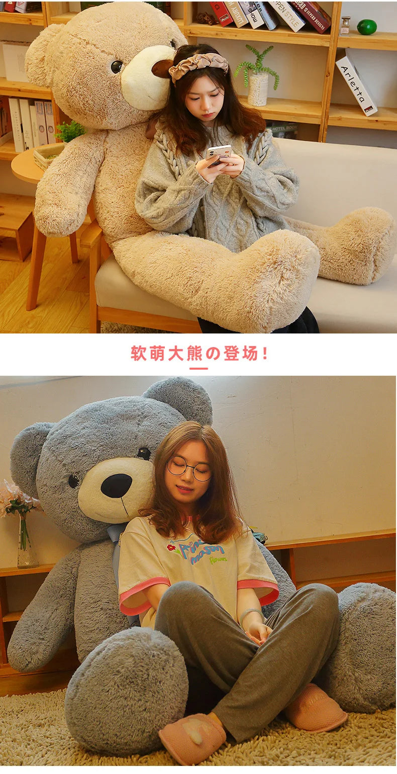 A young woman is sitting on a white sofa with a giant gray plush teddy bear, looking at her phone.