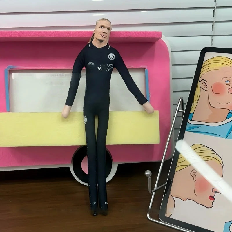A male football star stuffed doll wearing a black long-sleeved outfit standing in front of a pink display box.