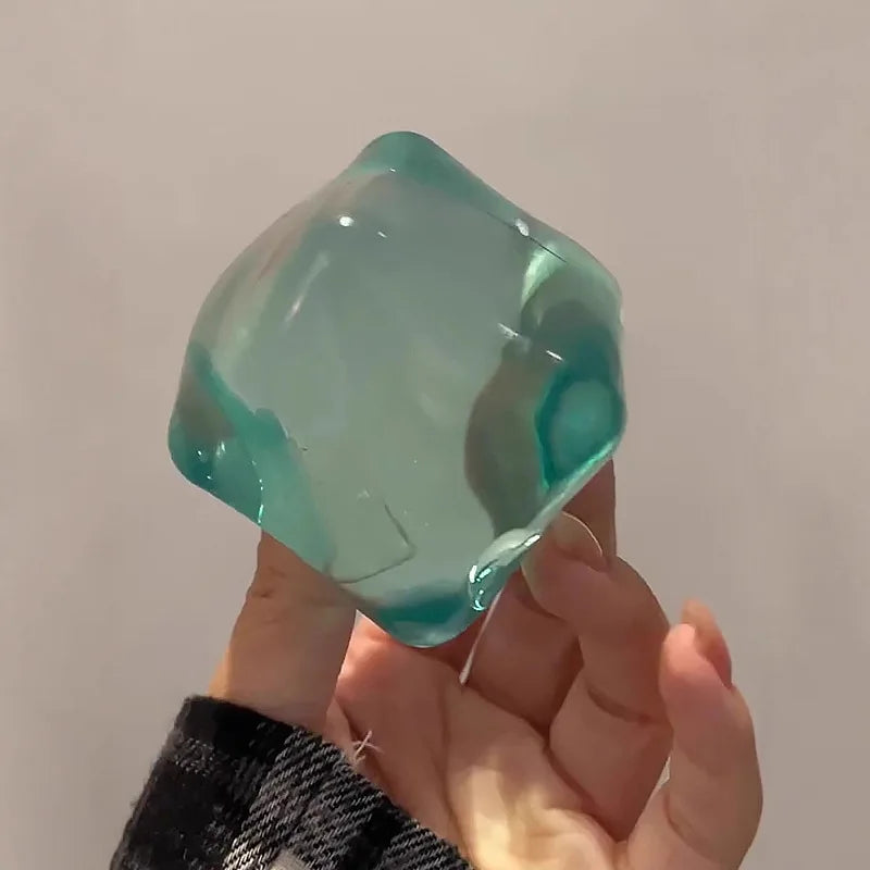 A hand holds a translucent teal ice cube sensory fidget toy against a light background.