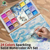 A watercolor paint set featuring 24 sparkling solid colors arranged in a tray, with a hand applying paint to paper.