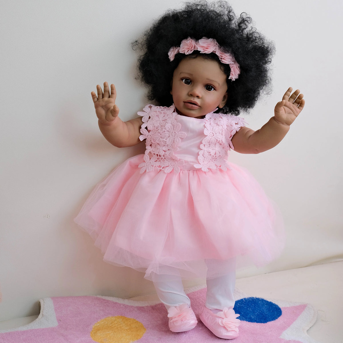 A pink reborn toddler doll with hand-rooted black hair wearing a pink dress standing on a colorful mat.