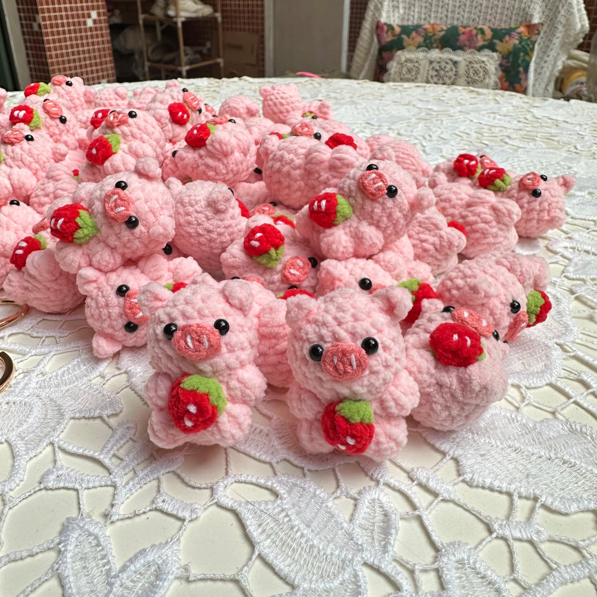 A pile of handmade crochet mini pig keychains in pink and red are displayed on a white lace tablecloth.