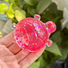 A hand holds a translucent pink squishy star turtle fidget ball with glitter accents against a blurred green background.