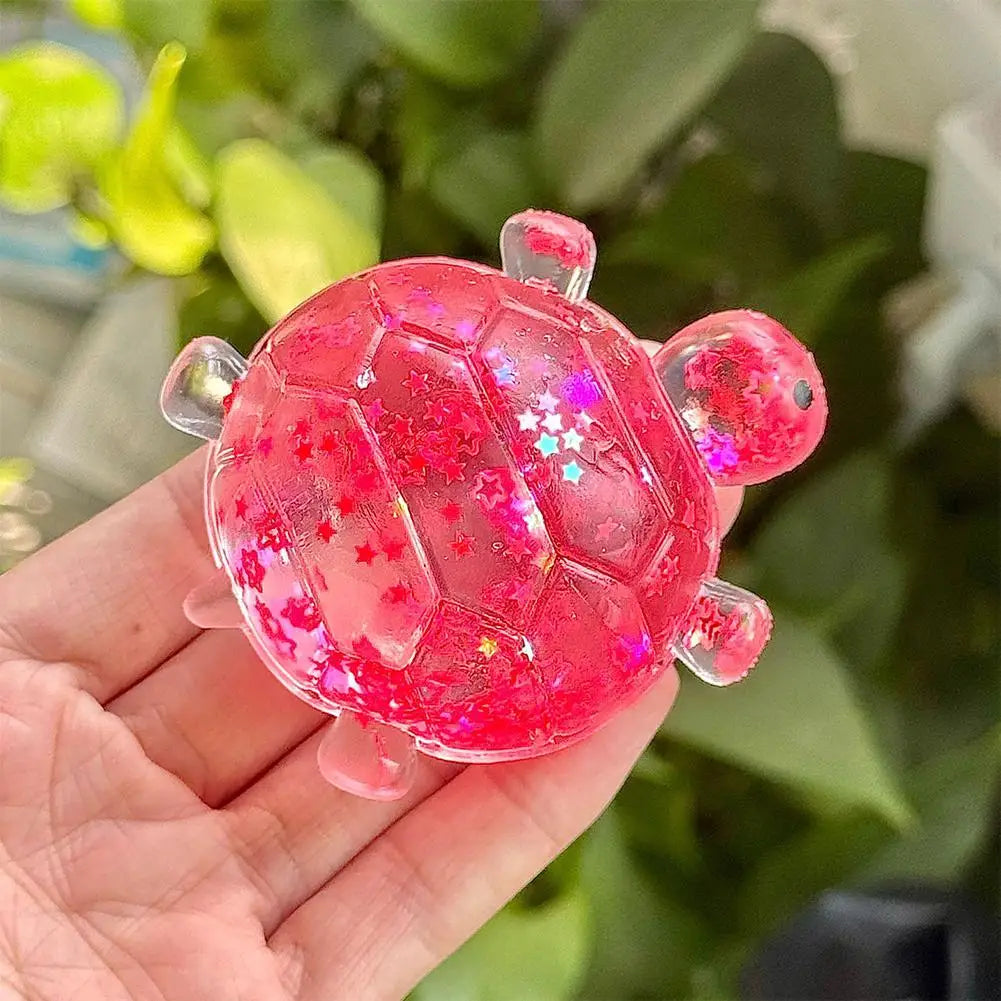 A hand holds a translucent pink squishy star turtle fidget ball with glitter accents against a blurred green background.