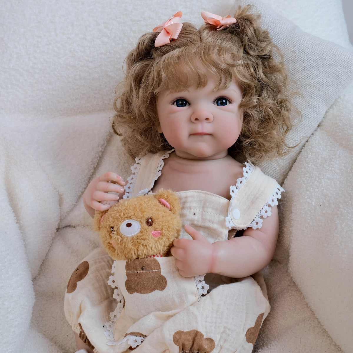 A baby doll with hand-rooted curly hair wearing a light-colored dress and holding a teddy bear.