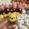 A hand holds a yellow crocheted animal keychain among various pink, brown, and white mini animals.