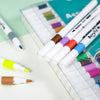 A collection of acrylic art markers in various colors is displayed in a plastic case.